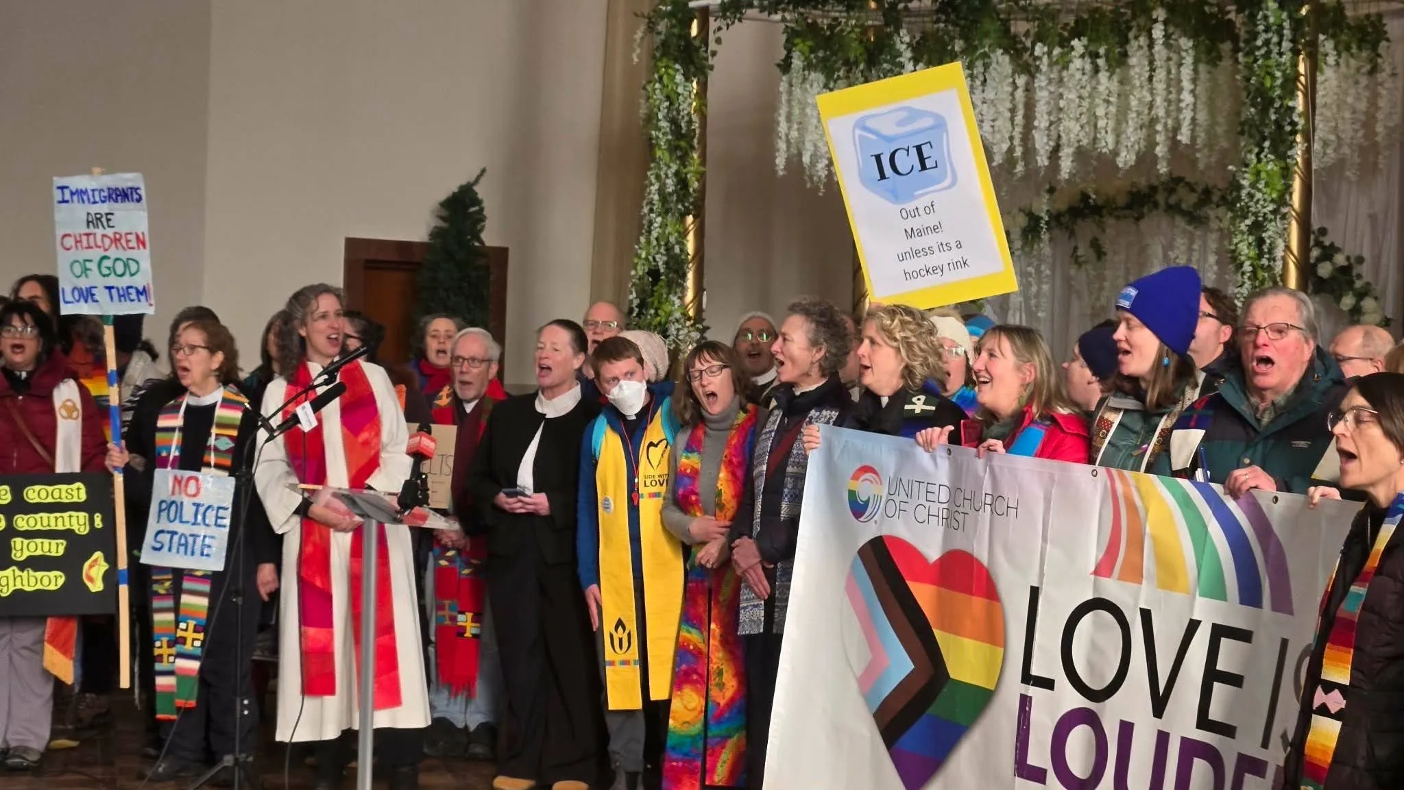 Clergy in bright stoles sing at an interfaith rally beneath a ‘Love is Louder’ Progress-Pride banner, with a sign reading ‘Immigrants are children of God’