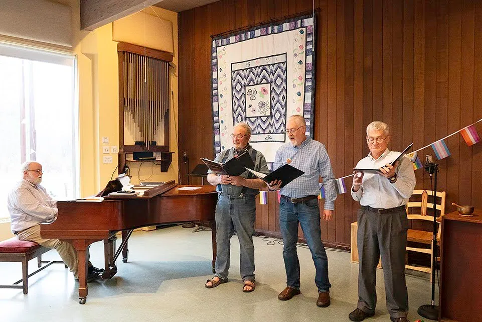Members make music together at the piano, singing from folders, with Pride and transgender flags strung behind them