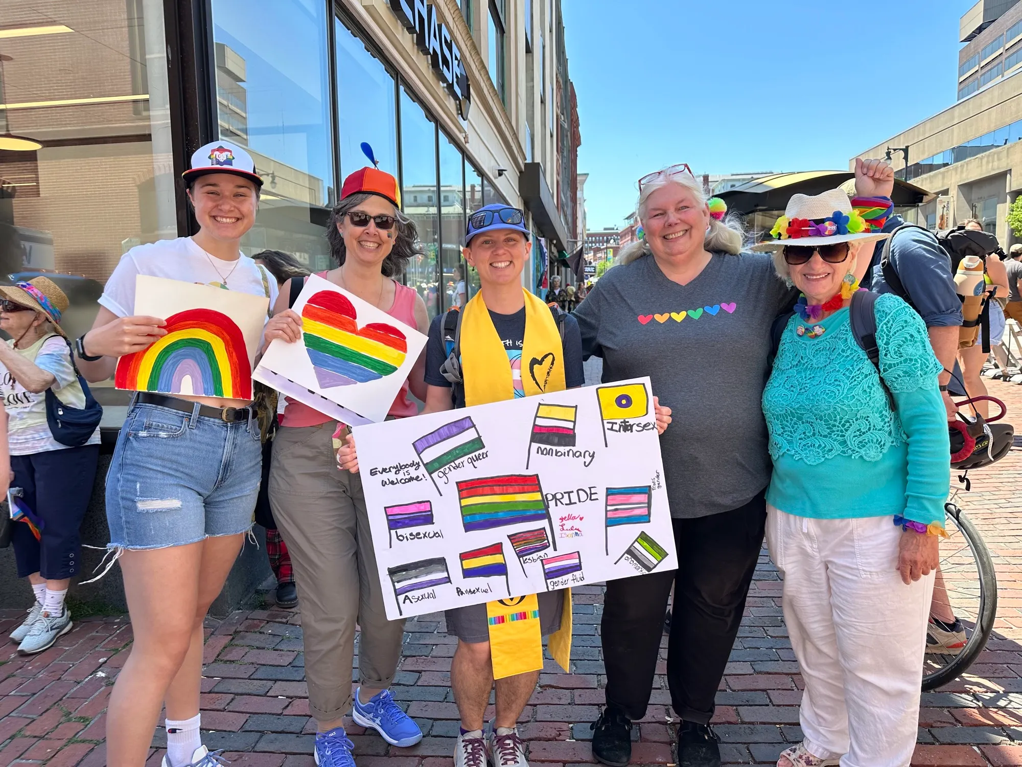 Five members pose at Portland Pride with hand-drawn rainbow hearts and a poster of identity flags