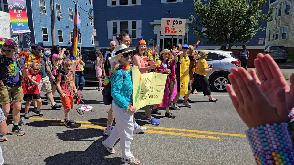 Members carry a Unitarian Universalist banner and a hand-made ‘Proud of all of U’ sign in a Pride parade, children waving pinwheels alongside