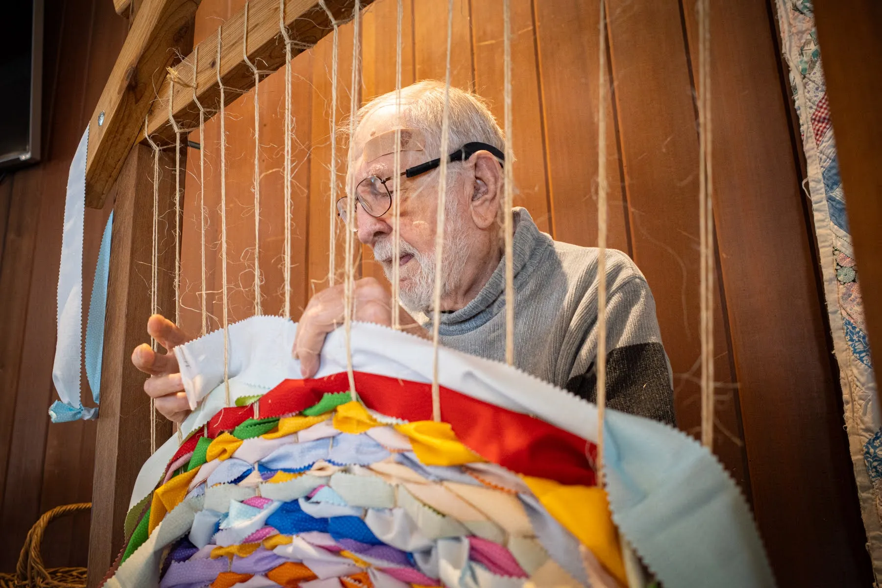 An older member hand-ties brightly colored fabric strips into a large communal weaving in a wood-paneled room