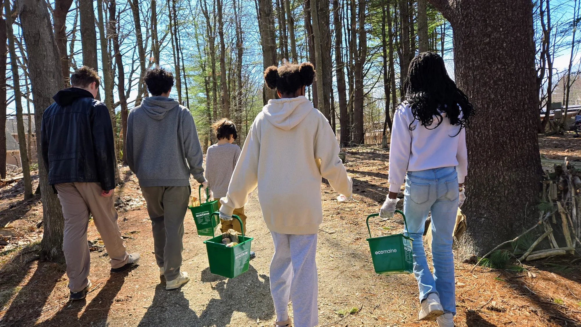Five teenagers walk a wooded path carrying sap buckets and wearing gardening gloves on a bright spring day